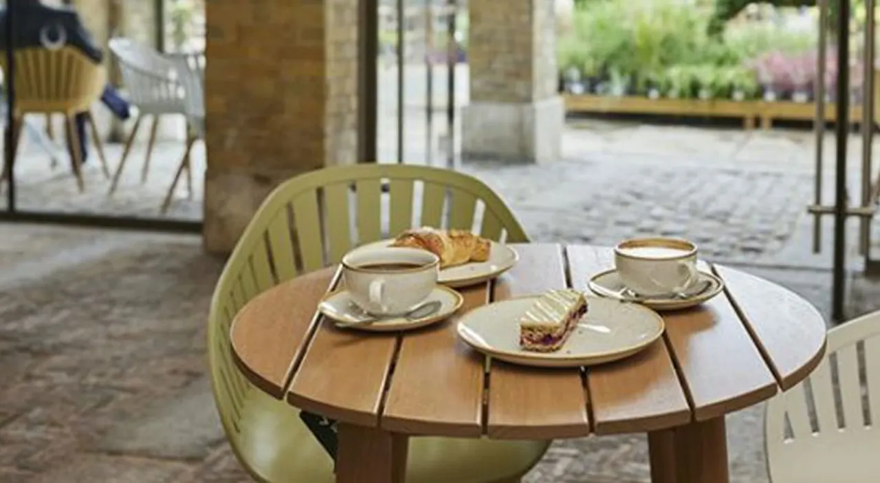 Two cups of coffee and pastries on a round wooden table set for two in a covered outdoor courtyard with stone pillars.