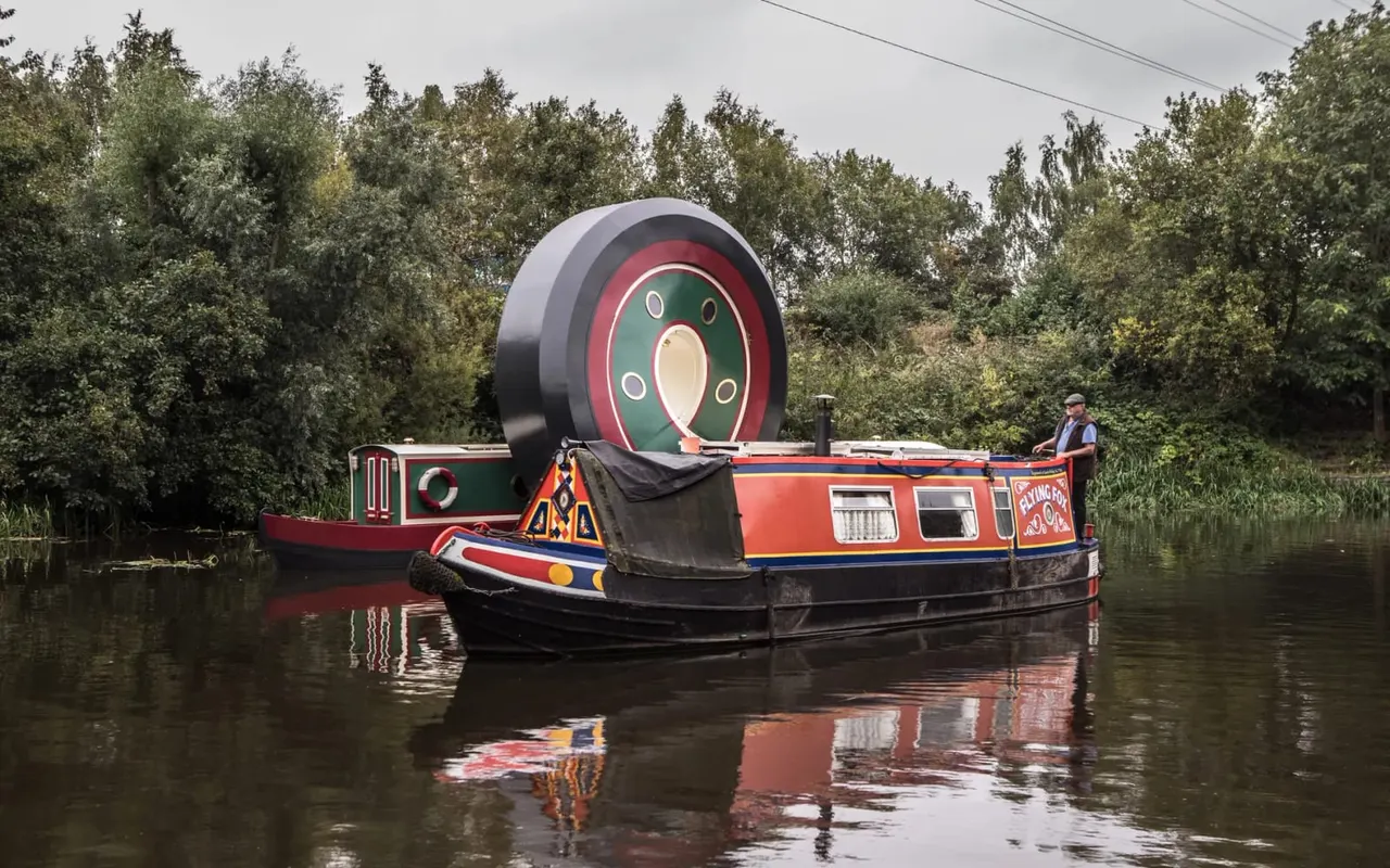 Brightly painted narrowboat moored on a canal with a large decorative wheel mounted on its roof and trees in the background.