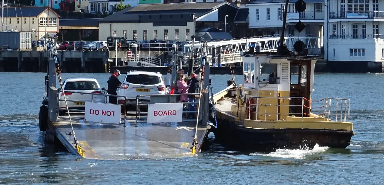 A car ferry carrying vehicles and passengers approaches the shore at a coastal town dock with colourful waterfront buildings in the background.