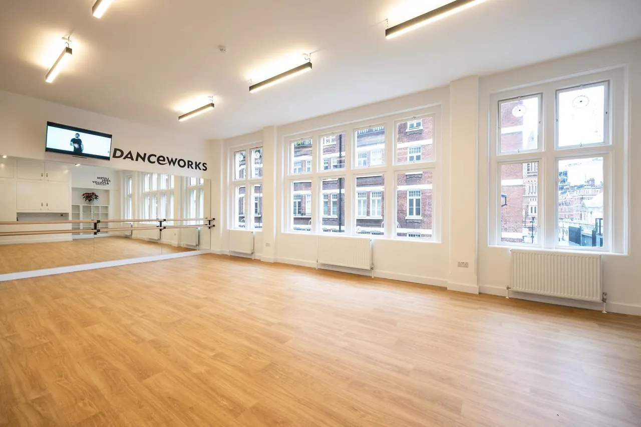 Empty dance studio with wooden flooring, wall mirrors and ballet barres, large windows overlooking a city street.