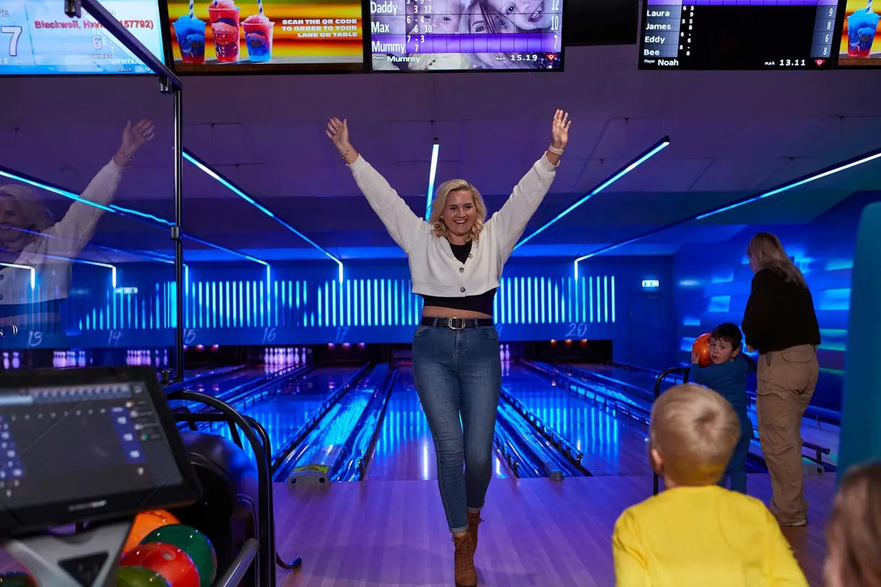 Woman celebrating with arms raised at a bowling alley with glowing neon lanes and other visitors nearby.