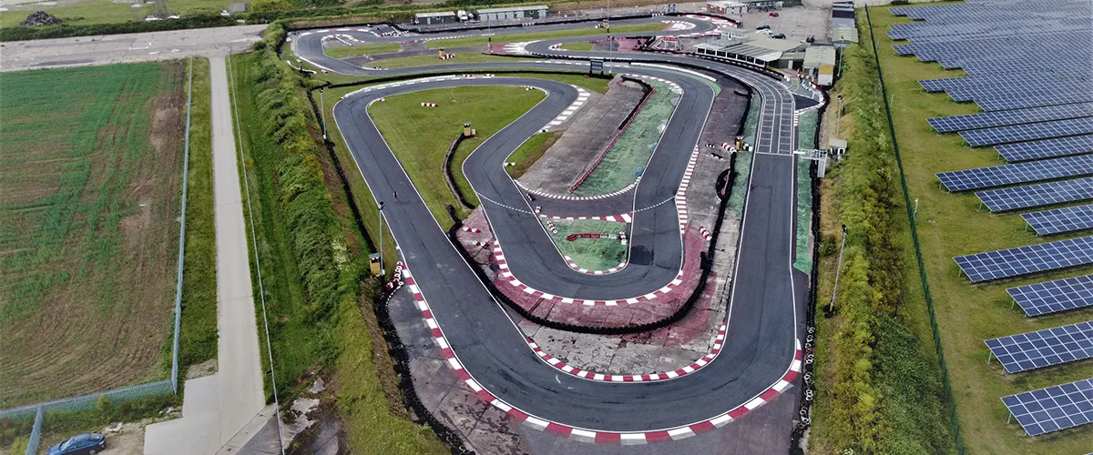 Aerial view of a kart racing circuit with multiple tracks, grandstands, and solar panels beside farmland.