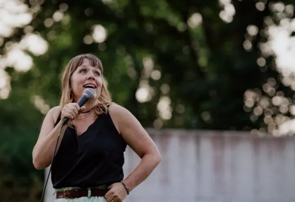 Woman holding a microphone, speaking outdoors with trees in the background.