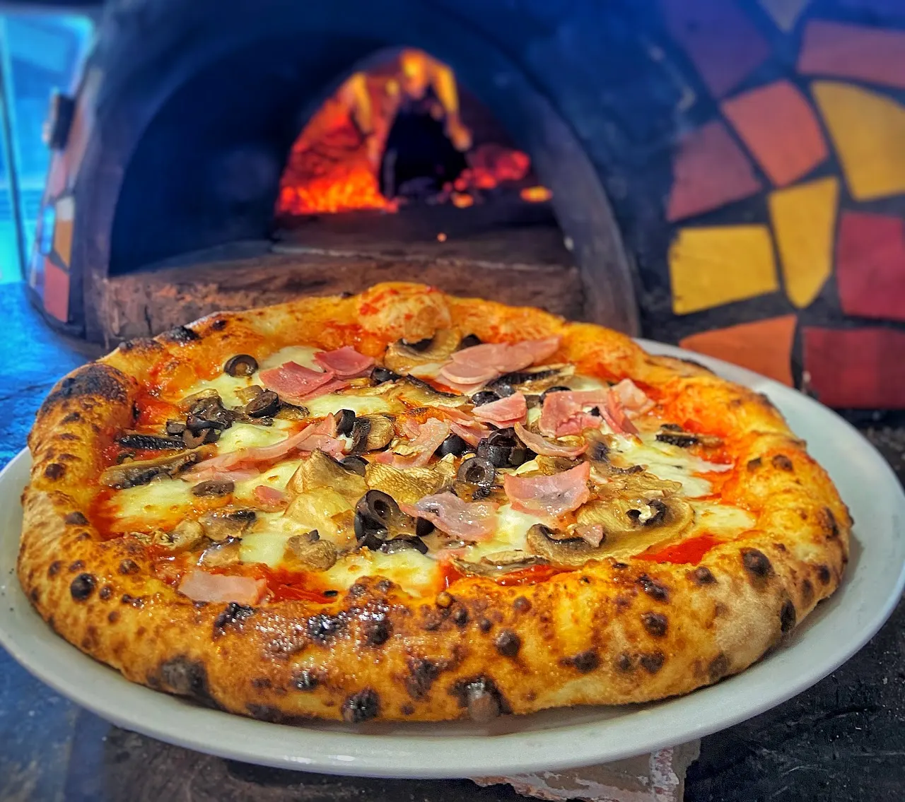 Wood-fired pizza topped with ham, mushrooms and olives, served in front of a lit pizza oven.