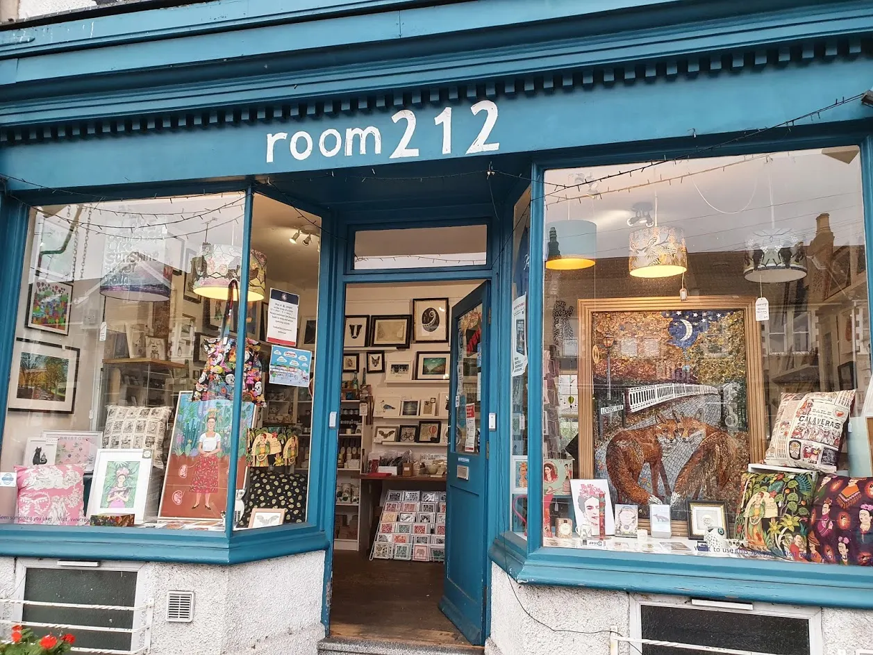 Blue-framed art gallery storefront with window displays of paintings, prints, and illustrations visible inside and on display shelves.