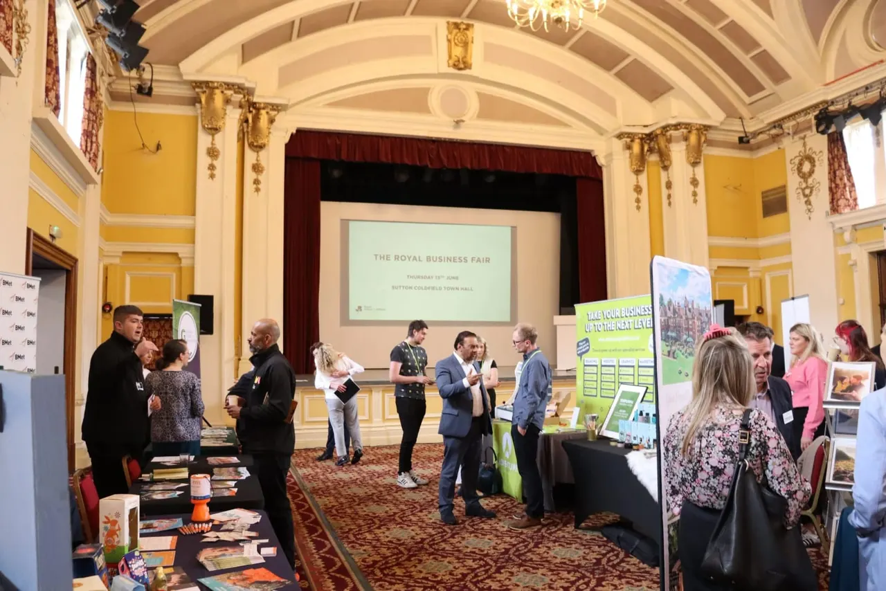 Attendees networking at The Royal Business Fair in an ornate hall with yellow walls, cream ceiling and a projection screen displaying the event title.