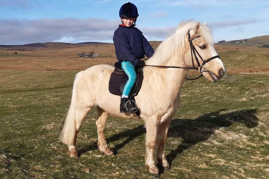 Child on a pale horse wearing a riding helmet, in an open moorland landscape with hills and farm buildings in the distance.