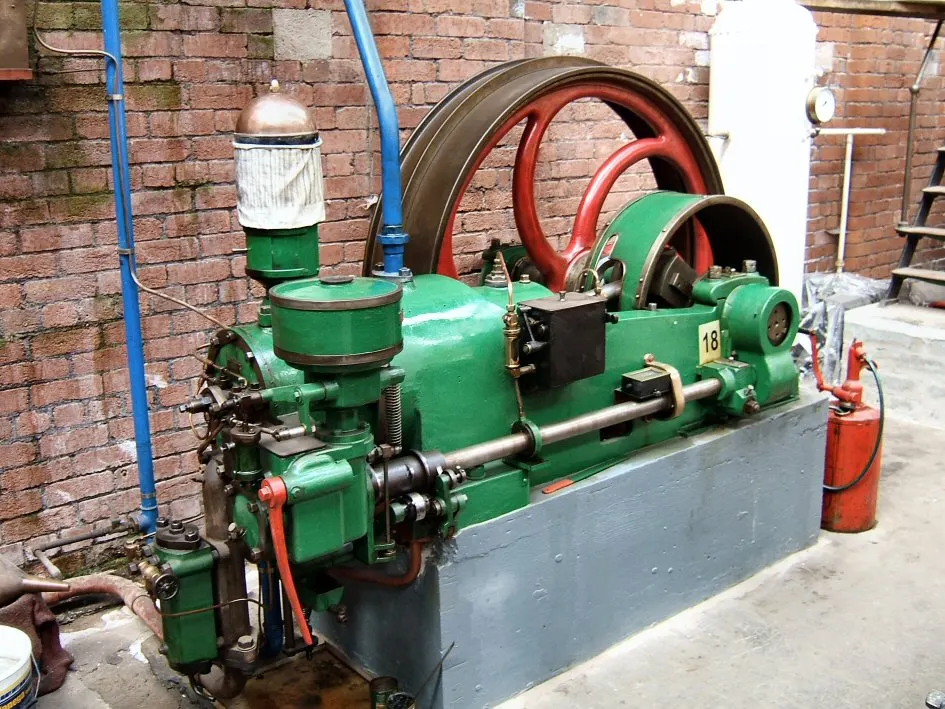 A vintage green steam engine displayed against a brick wall with red and blue piping.
