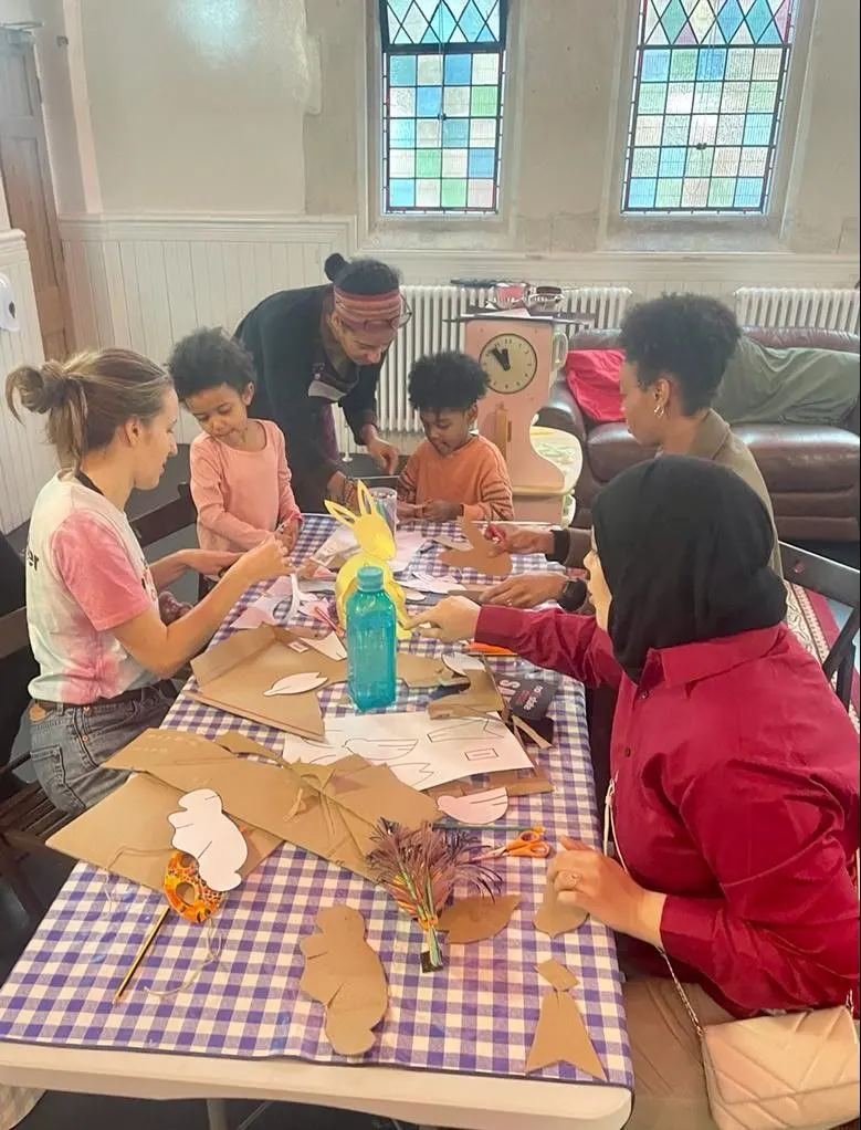 Children and adults doing a craft activity at a table with purple and white checked cloth in a community space.