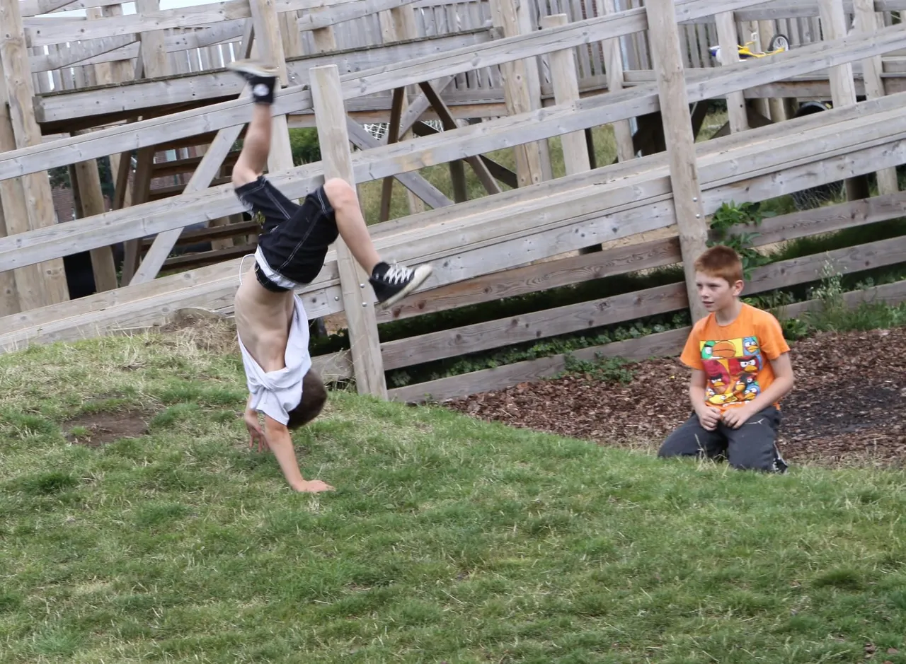 Child performing a handstand on grass while another child watches nearby, wooden fencing in background.