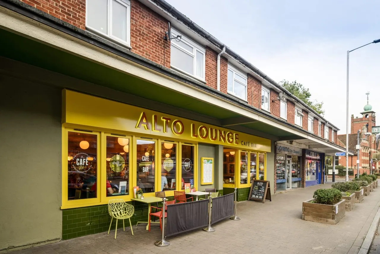 Bright yellow-fronted Alto Lounge café bar with outdoor seating on a town high street, featuring green and yellow décor and brick residential buildings above.