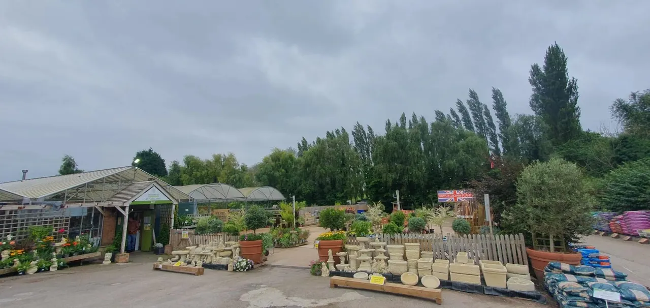 Garden centre with greenhouses, plant displays, paving stones and landscaping materials on sale, surrounded by tall trees.