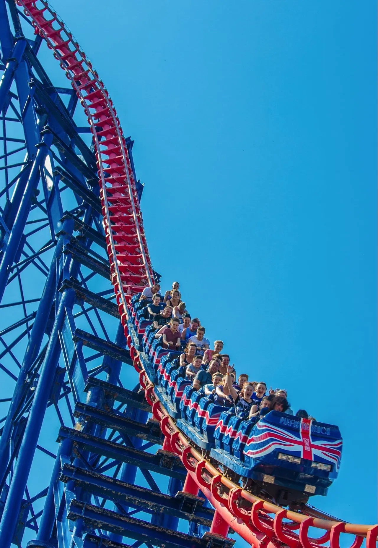 Roller coaster train full of riders in red seats climbing a steep blue track structure against a clear blue sky.