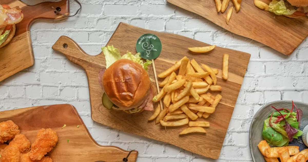 Burger with lettuce and ham served with chips on a wooden board against a white brick wall.