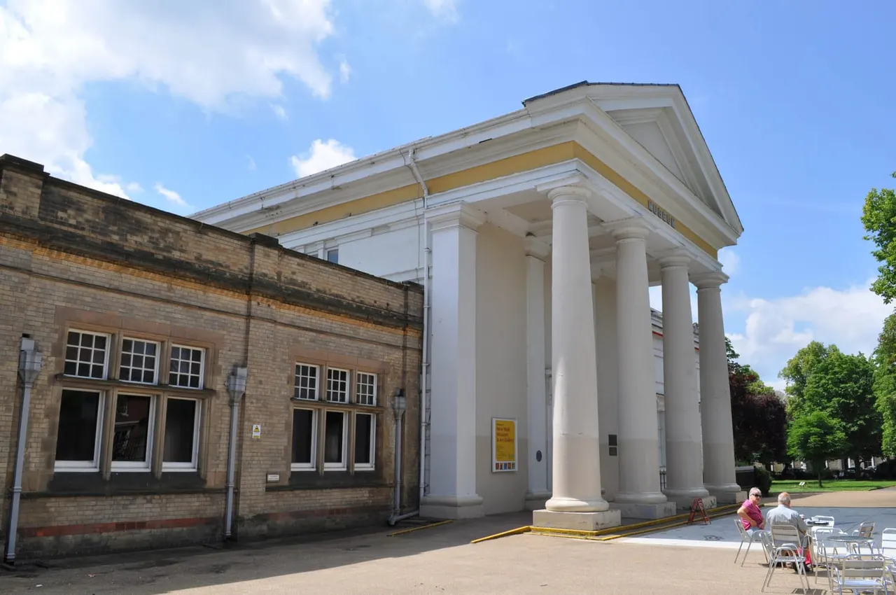 Classical building with white columns and yellow trim, seated visitors in foreground, adjacent brick building on left.