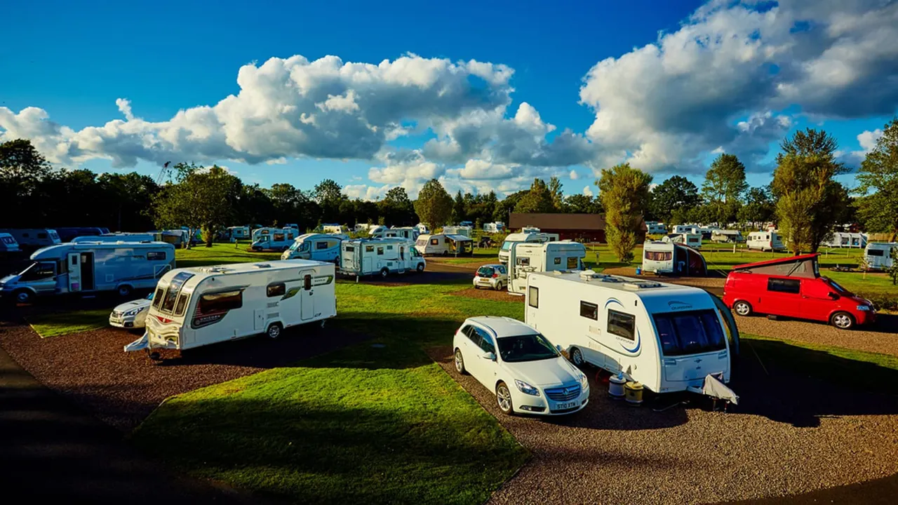 Caravan and motorhome campsite with numerous recreational vehicles parked on grass and gravel pitches under blue sky with white clouds.