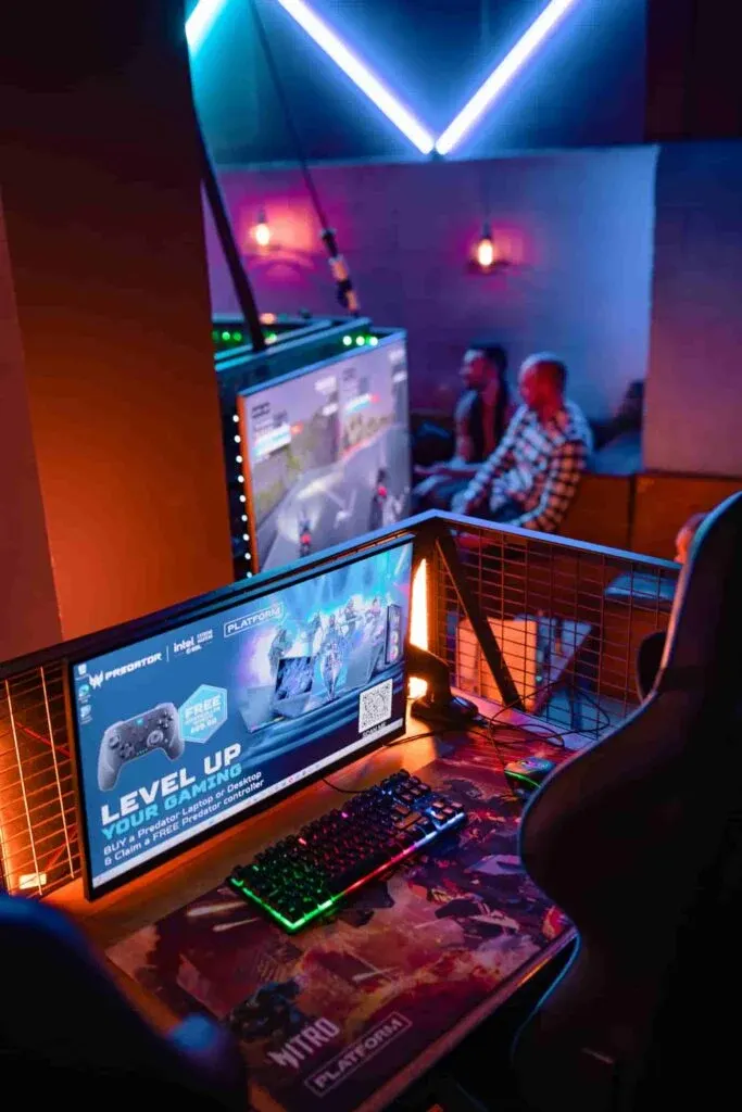 Gaming station with illuminated monitors and keyboard in a neon-lit arcade venue with players in background.