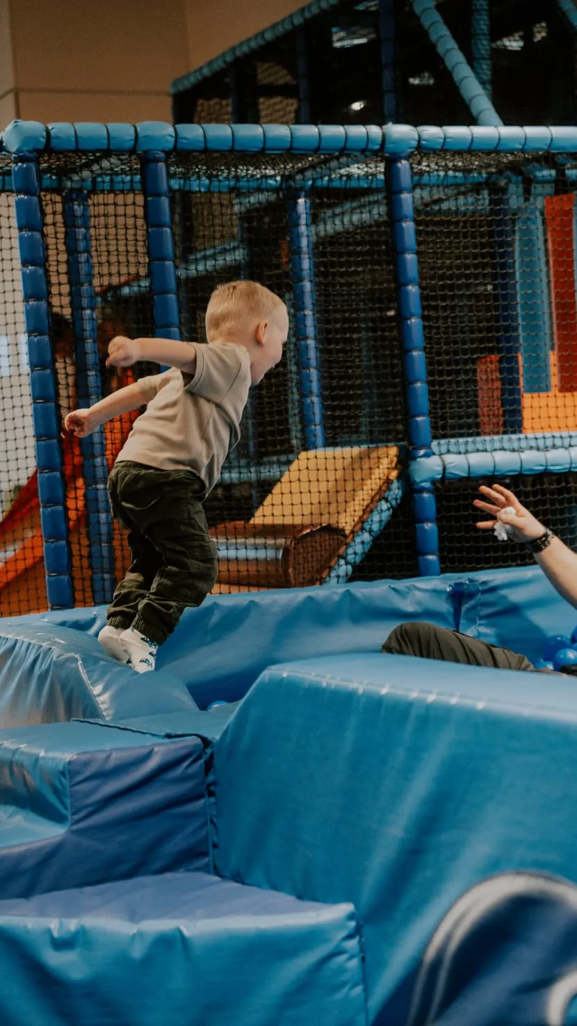 A toddler jumps on a blue inflatable play structure with netting and padding inside an indoor play centre.