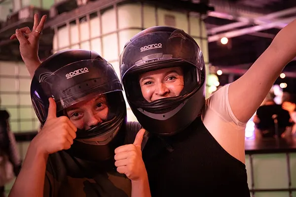 Two people wearing protective helmets give thumbs-up gestures inside an indoor entertainment venue with pink lighting.