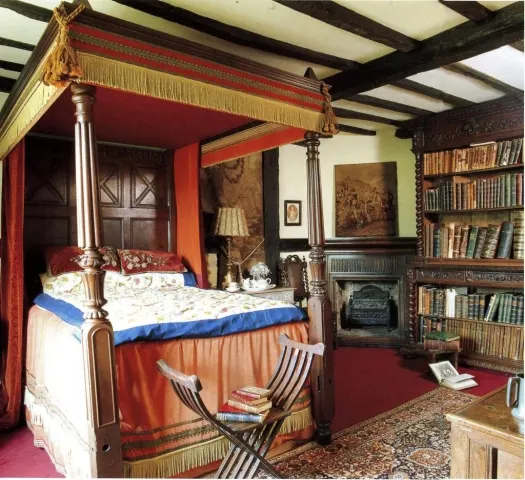 A luxurious four-poster bed with red and gold drapes in a period bedroom with bookshelves, fireplace and historical furnishings.