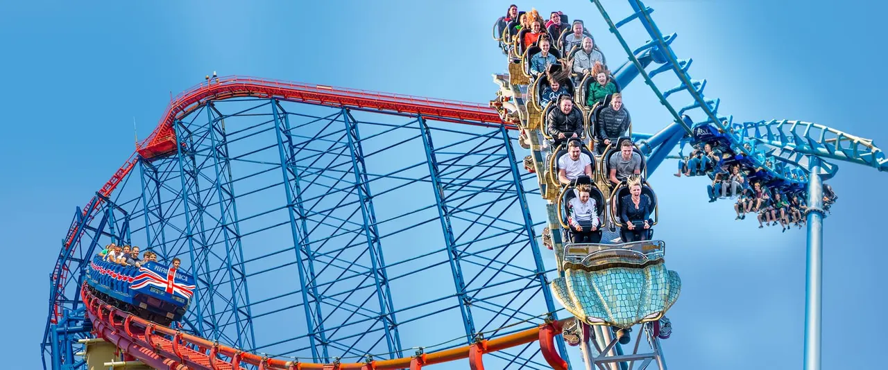 Visitors on two roller coaster rides against a clear blue sky at an amusement park.