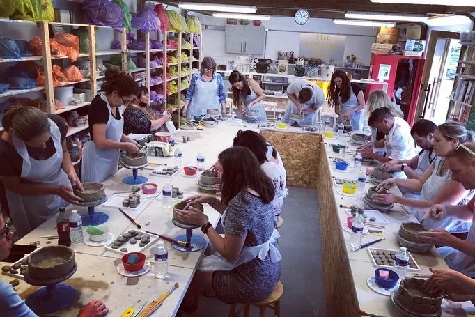 A group of people seated at long tables engaged in pottery painting or decorating activity in a bright studio space.