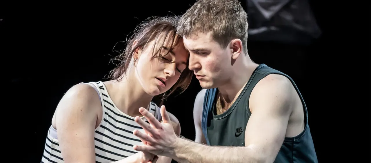 Two actors in an intimate scene during a stage performance, heads close together against a black background.