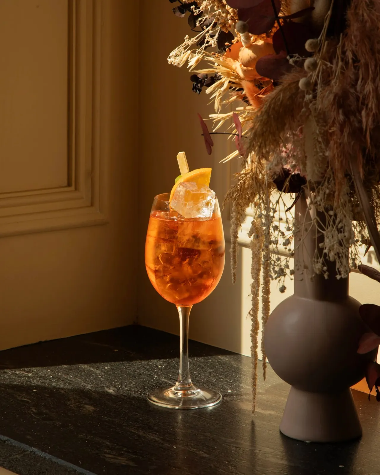 An orange cocktail with ice and orange slice garnish on a bar counter, styled with dried flowers and decorative elements.