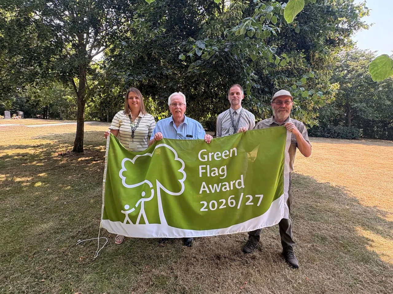 Four people holding a Green Flag Award banner in a woodland clearing surrounded by mature trees.