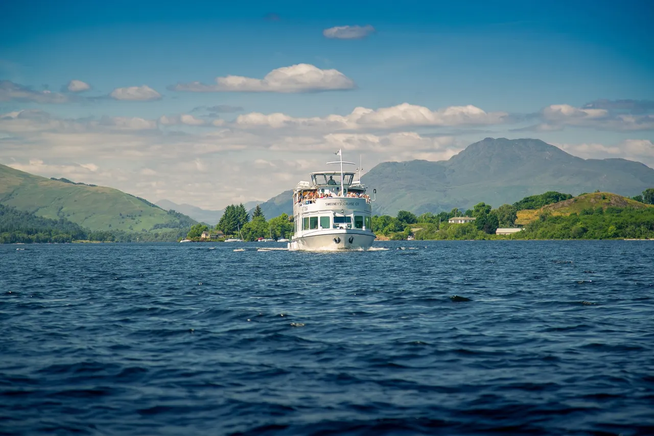 A white passenger cruise boat on Loch Lomond with mountains and green hills in the background under a blue sky with white clouds.