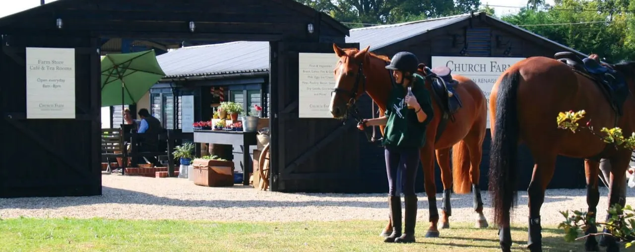 A rider in black helmet and jacket stands beside a chestnut horse on gravel near black farm buildings and a shop.