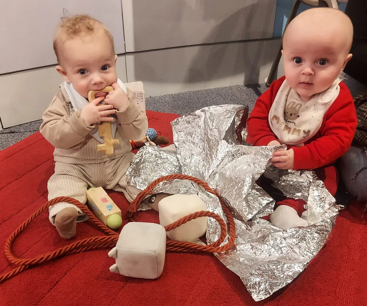 Two babies exploring sensory play items including wooden toys and crinkly silver material on a red carpet.