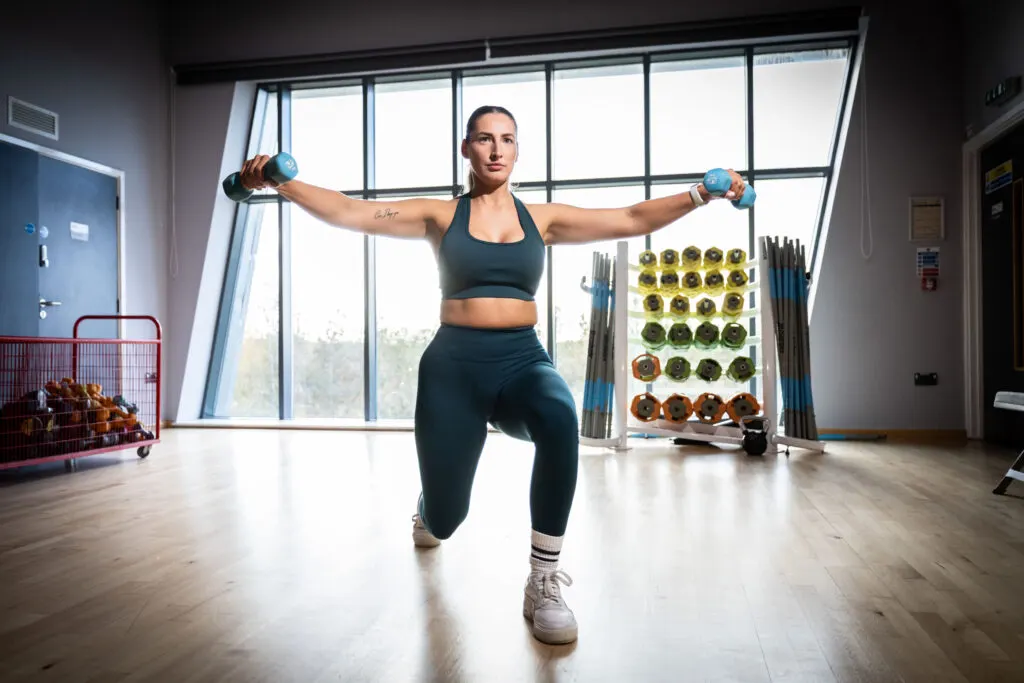 Woman performing a weighted lunge exercise with dumbbells in a modern gym studio.