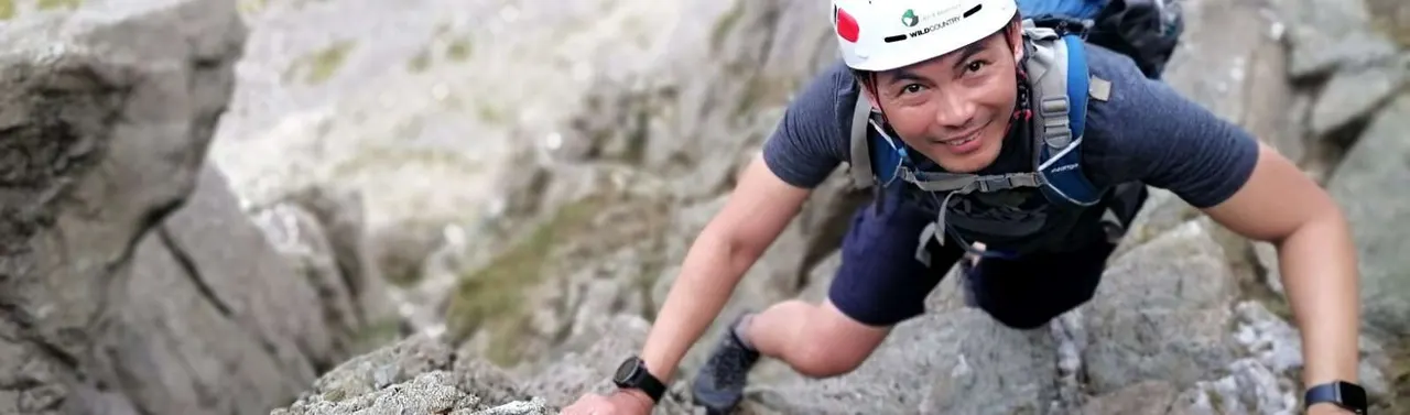 Person in climbing helmet and harness ascending rocky cliff face outdoors.
