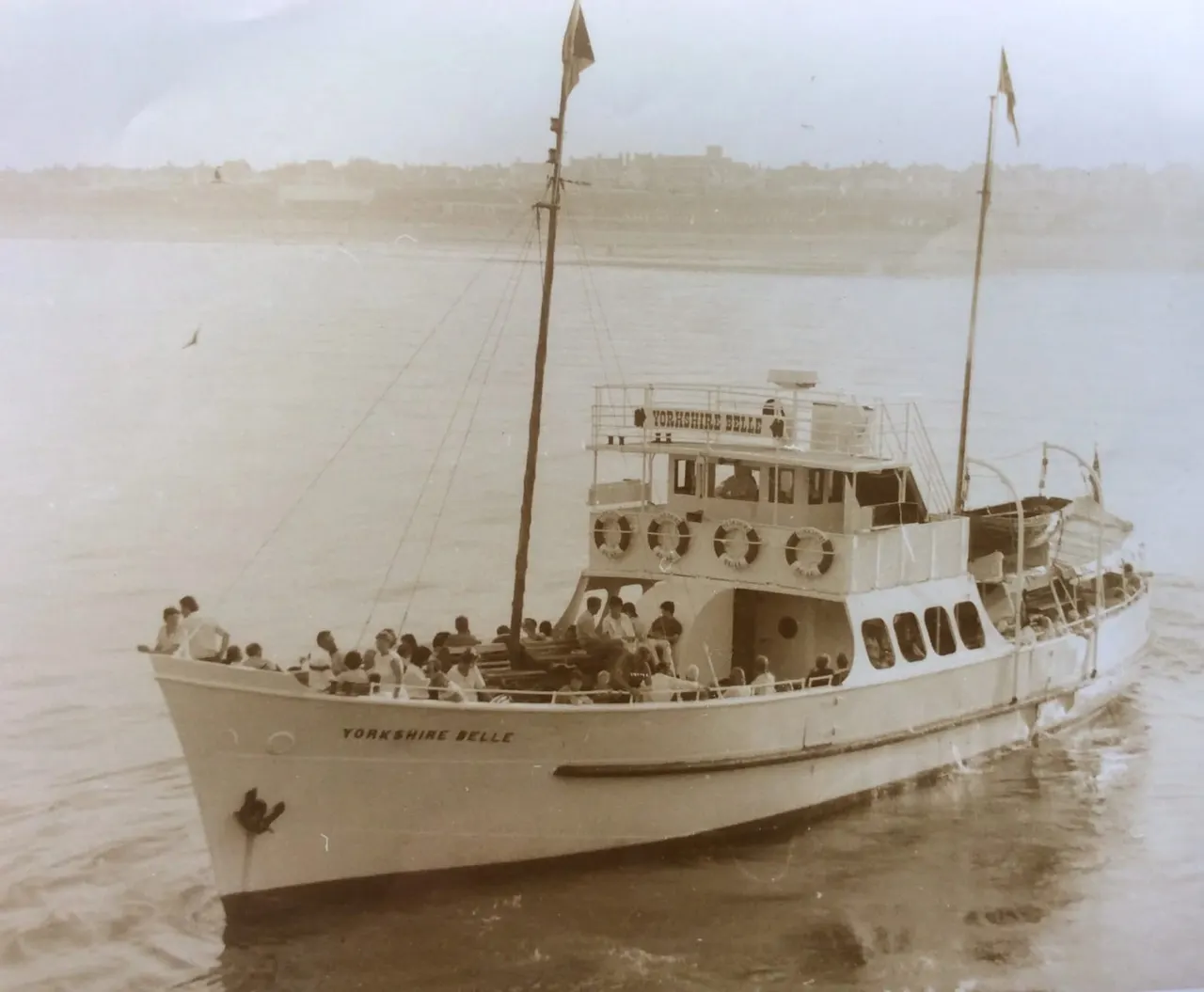 Vintage photograph of the Yorkshire Belle paddle steamer moored in shallow water with passengers aboard, coastal town visible in misty background.