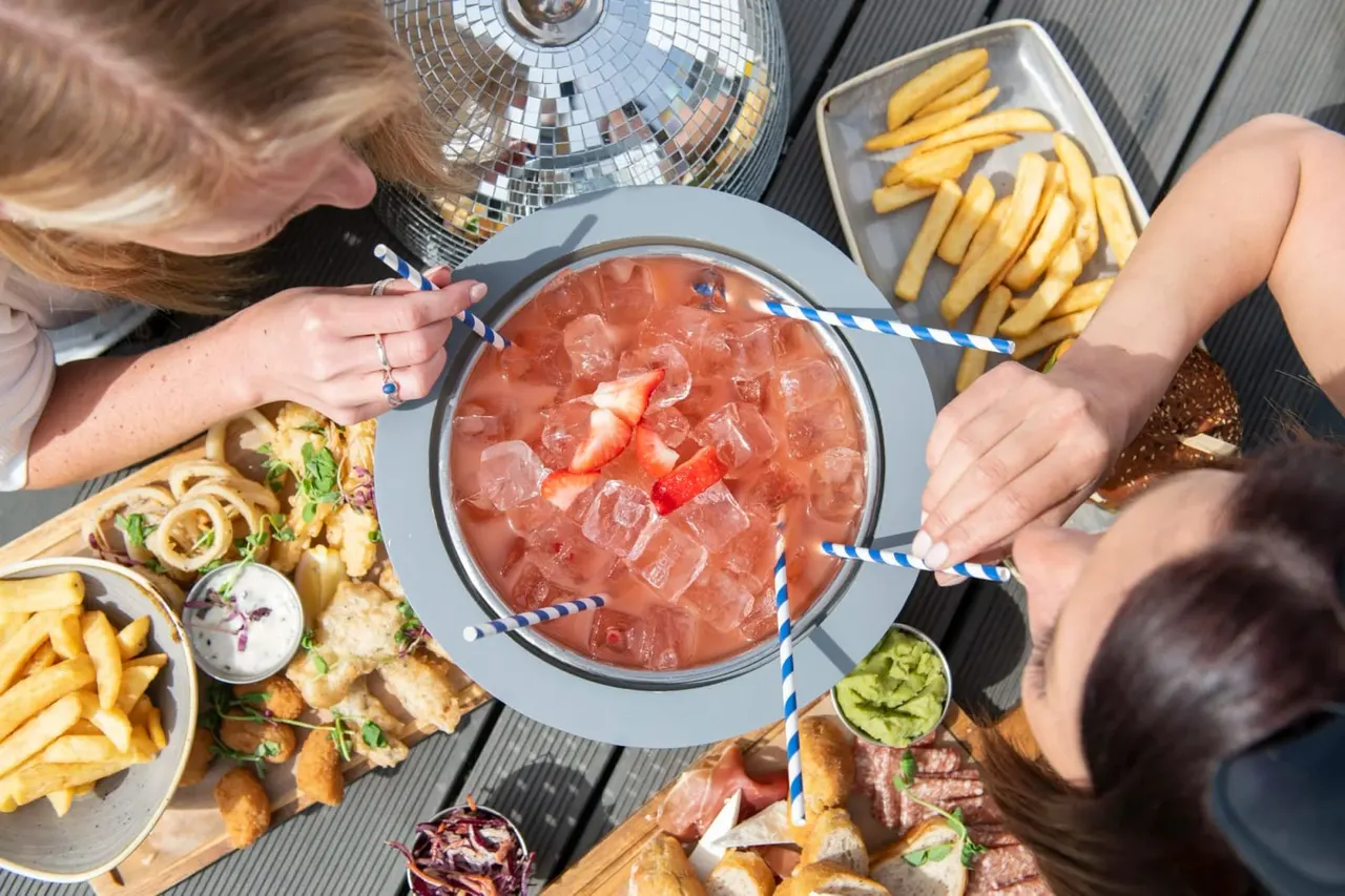 Two people sharing a bowl of strawberry cocktail with striped straws, surrounded by chips, cured meats, and other appetisers on a sunny outdoor table.