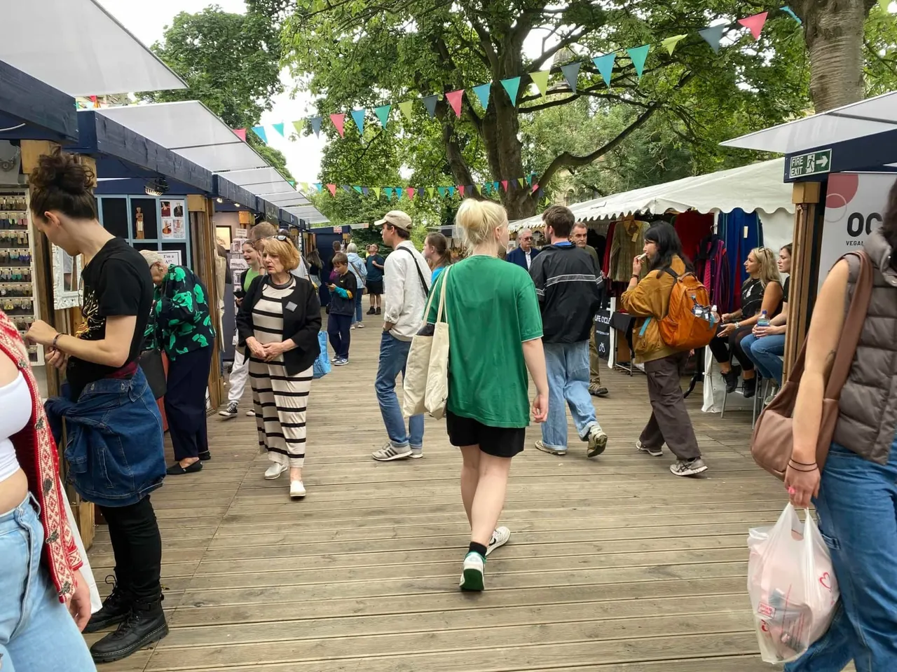 Outdoor market with visitors browsing stalls under bunting and shade structures, with large trees overhead.