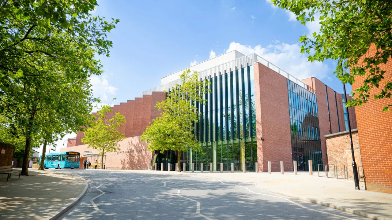 Modern brick and glass building with green trees framing the entrance and a blue bus parked outside on a sunny day.
