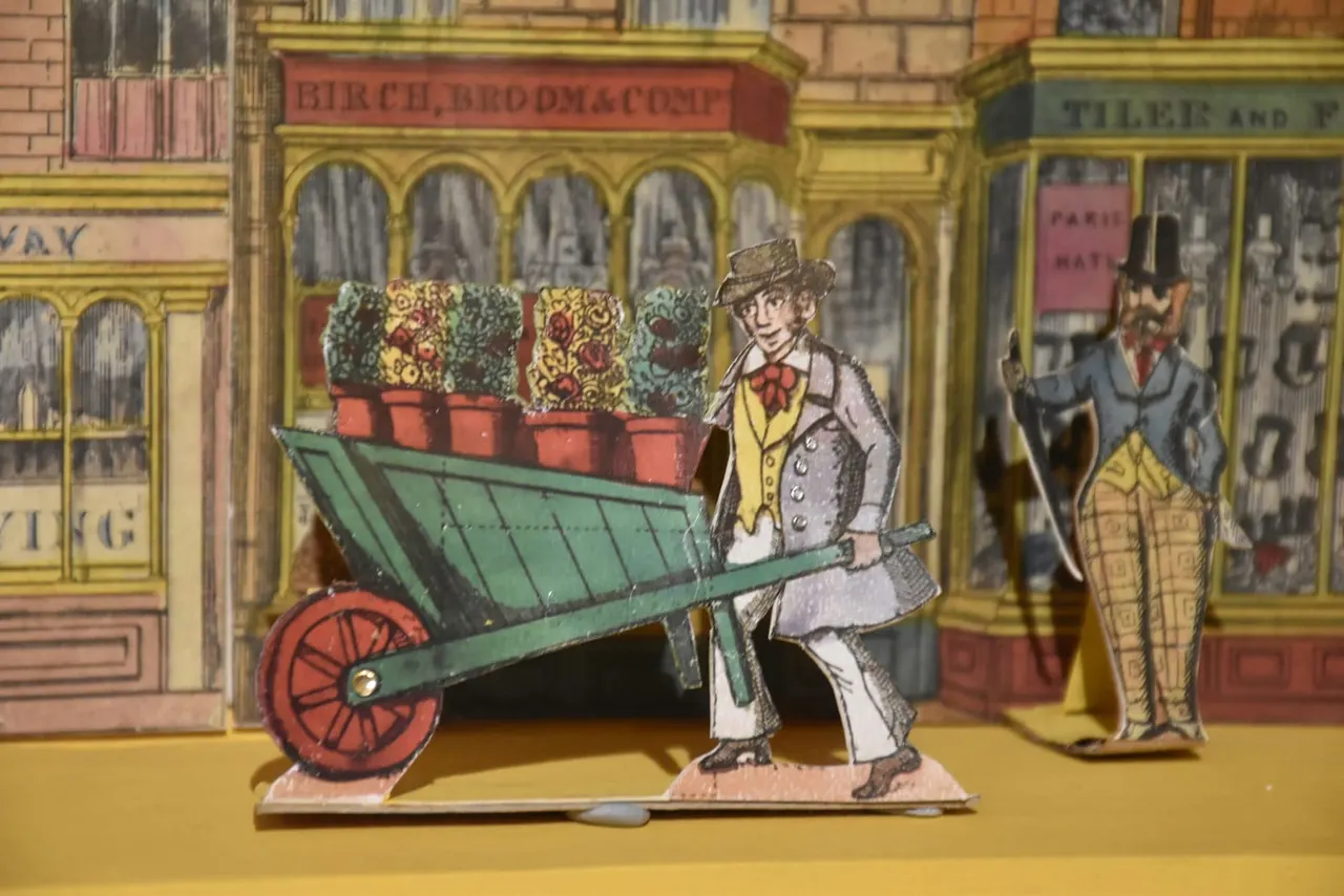 Hand-coloured paper theatre scene depicting a Victorian street with a figure pushing a wheelbarrow of flowers outside shops.