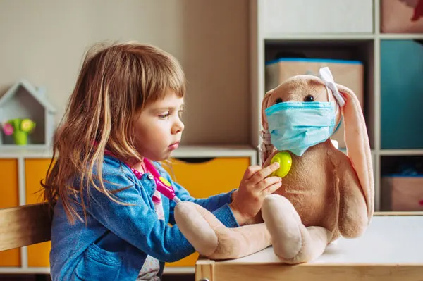 Young child in denim shirt playing doctor with a stuffed toy wearing a face mask.