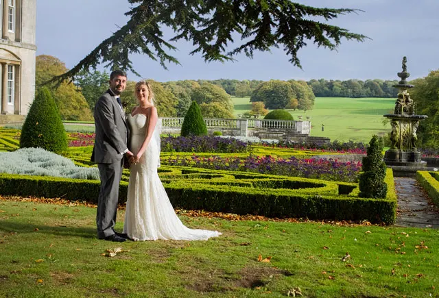 Bride and groom posing in formal gardens with manicured hedges, floral beds and country estate in background.