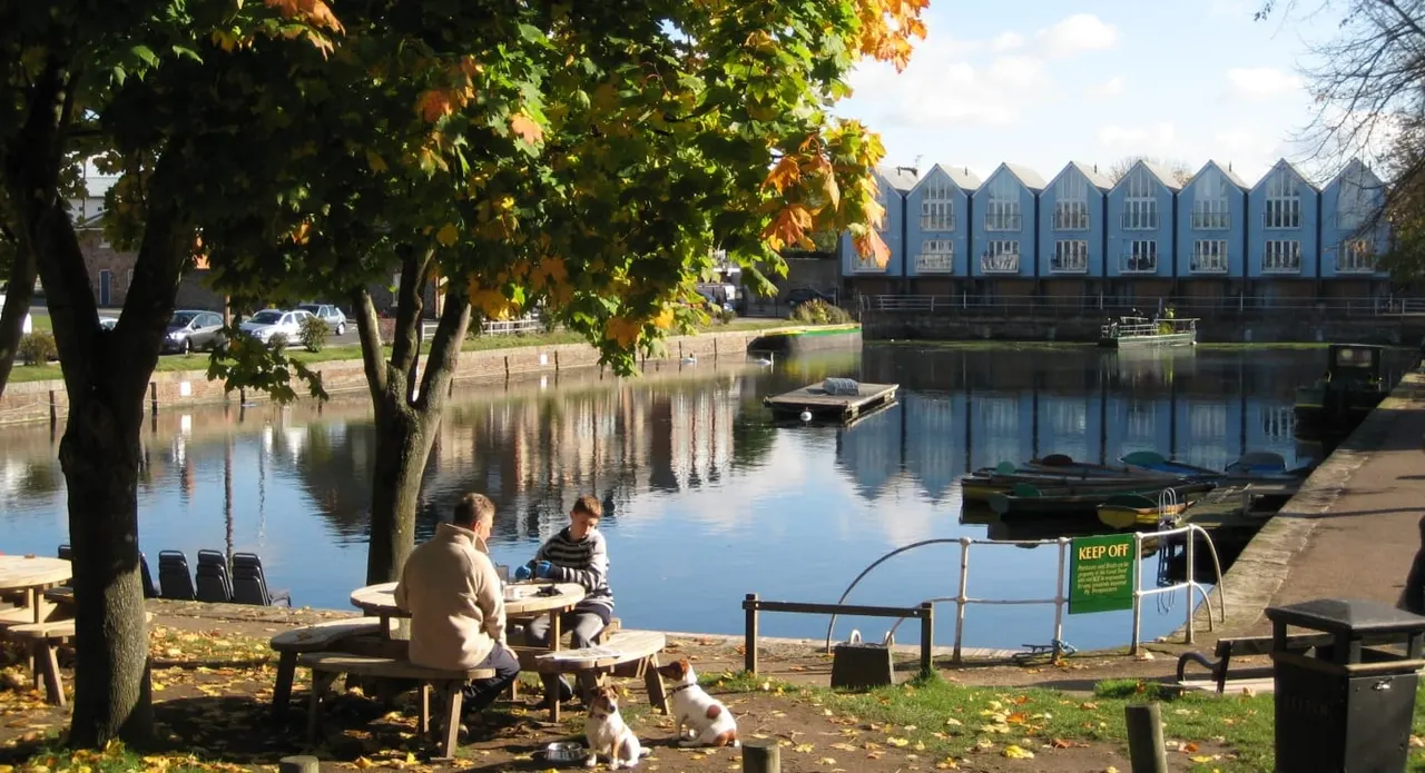 Two people and a dog sit at a picnic table by an autumn tree on the bank of a calm canal, with blue boathouses reflected in the water beyond.