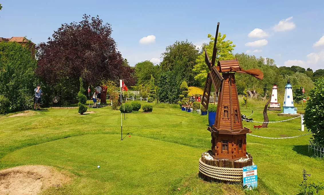A mini golf course with decorative wooden windmill and lighthouse structures on a green lawn, with visitors playing in the background on a sunny day.
