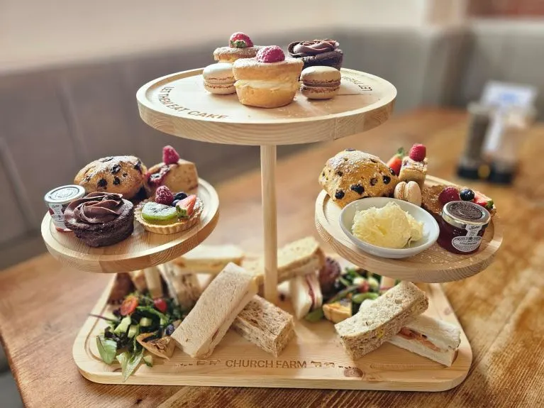 A tiered wooden stand displays an afternoon tea spread including cakes, pastries, scones, cream, jam, and finger sandwiches.