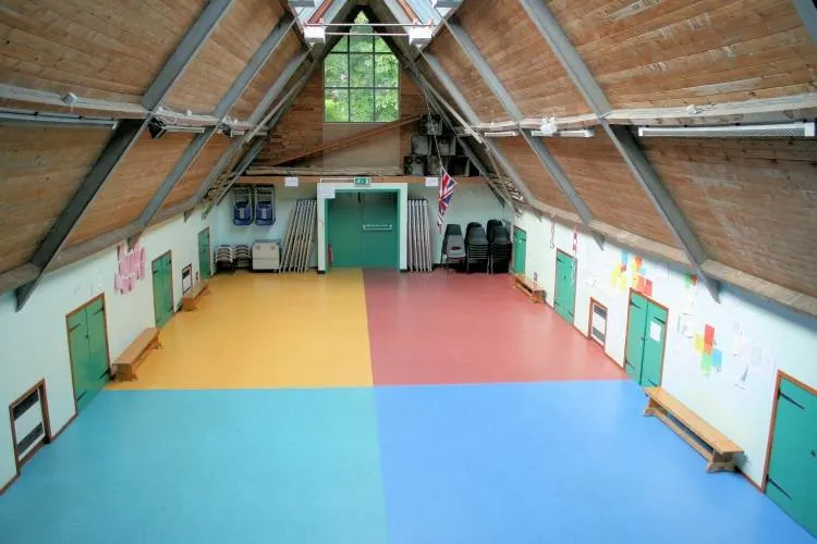 Interior of a scout hall with coloured floor sections, exposed wooden roof beams and green storage cupboards along the walls.