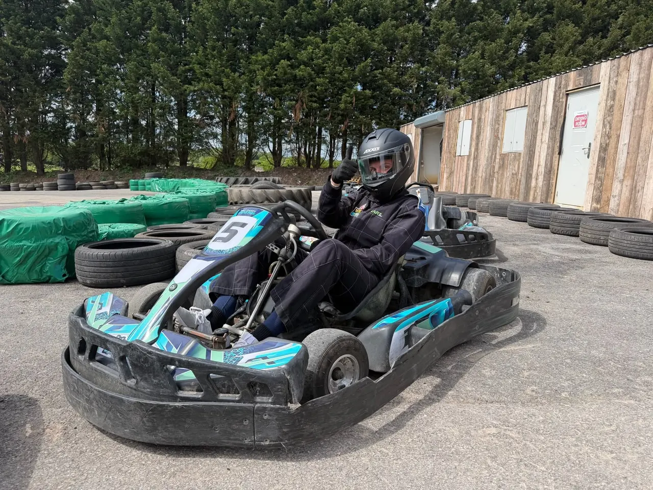 Person in helmet and black race suit driving a go-kart on an outdoor track lined with tyres.