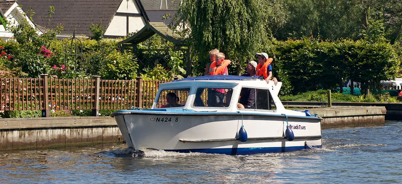 Passenger boat with children wearing orange life jackets on the roof, moving through a river past gardens and moored boats.