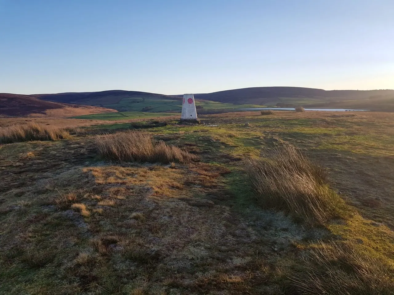 A white trig point stands on rolling moorland with hills and water visible in the distance under a clear sky.