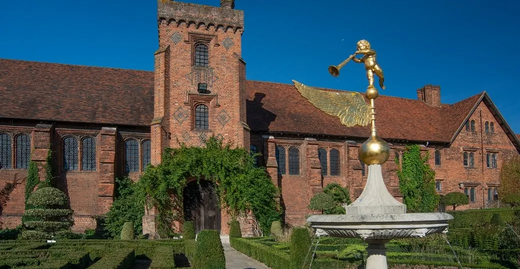 Red brick Tudor mansion with tower and gold statue of a figure with trumpet on a white stone pedestal, surrounded by formal gardens with topiary under clear blue sky.