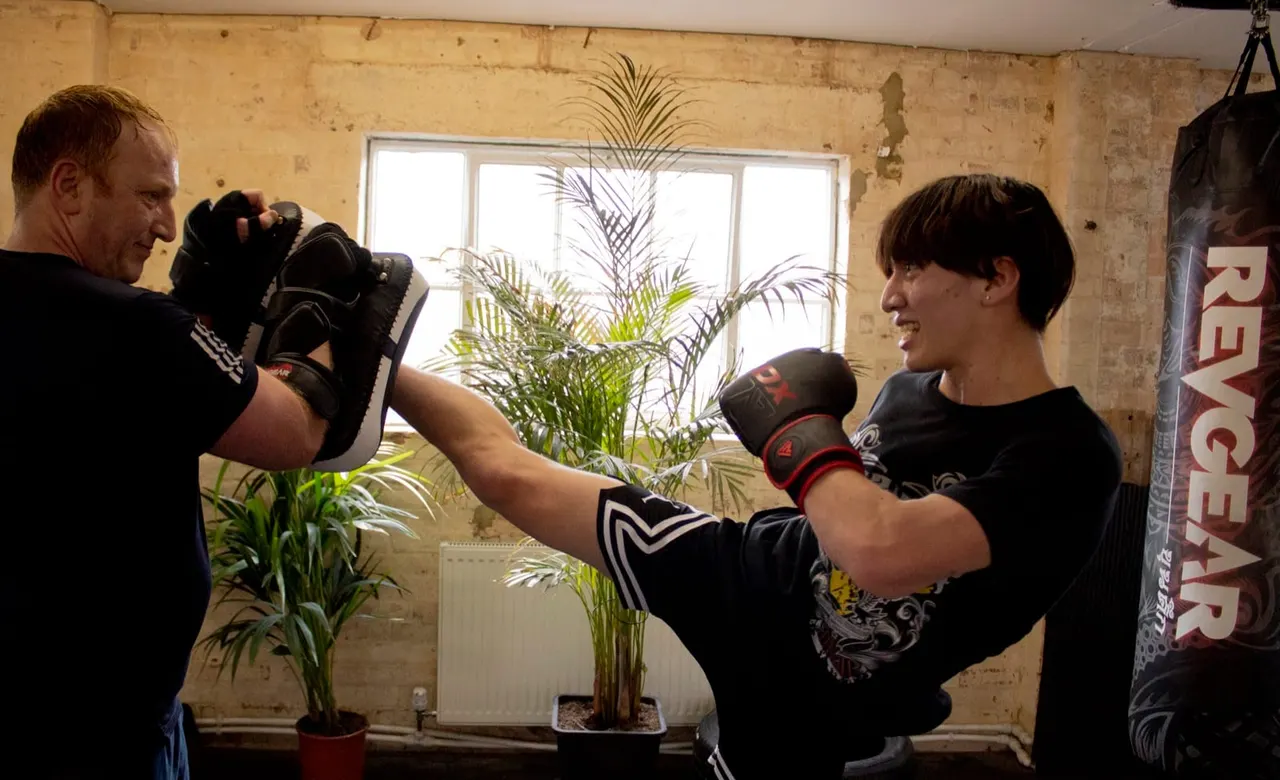 Two people boxing together indoors, one wearing black gloves throwing a punch while the other holds pads, in a gym with exposed brick walls.
